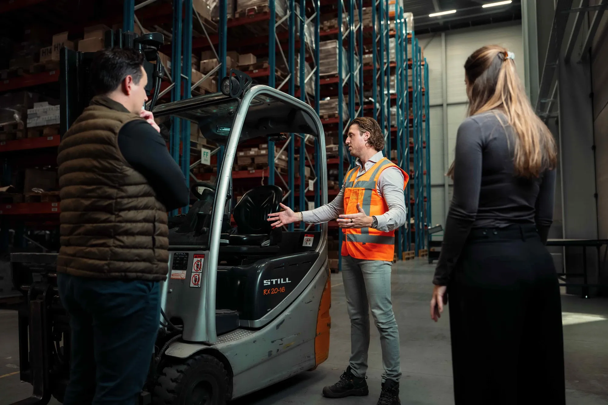 Man in een oranje veiligheidshesje geeft uitleg bij een heftruck in een magazijn aan twee toehoorders