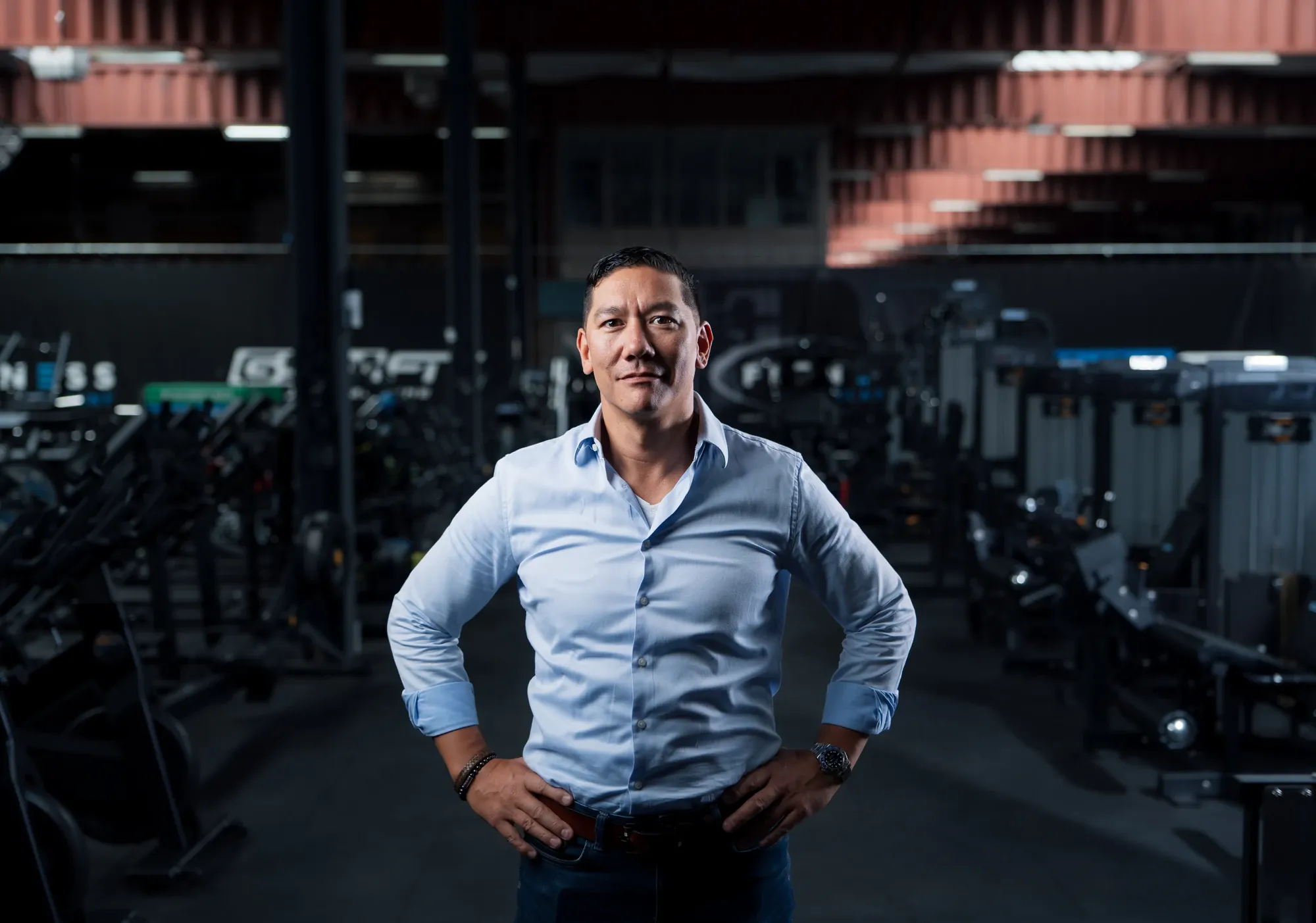 Man in een lichtblauw overhemd staat met de handen in de zij tussen de toestellen in de showroom van Fitness Company