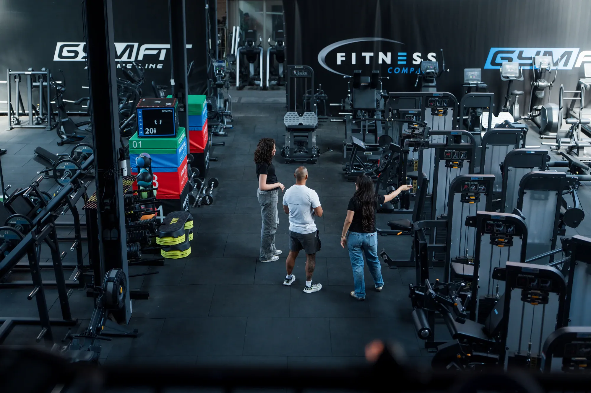 Bovenaanzicht van de showroom van Fitness Company, waar twee medewerkers een bezoeker langs de krachttoestellen begeleiden