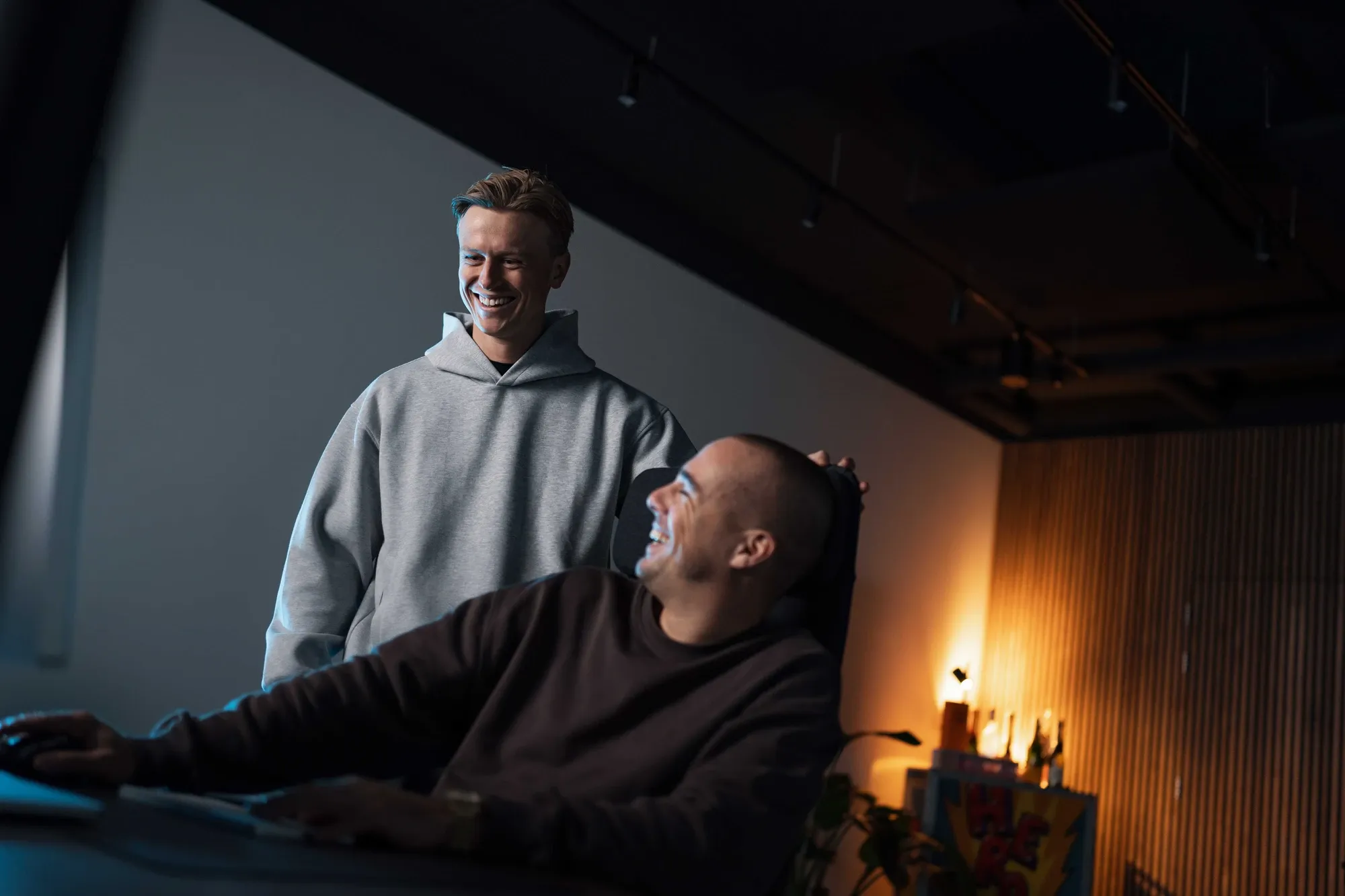 Twee collega’s van Klubb lachen samen bij een bureau, een van hen staand achter de ander