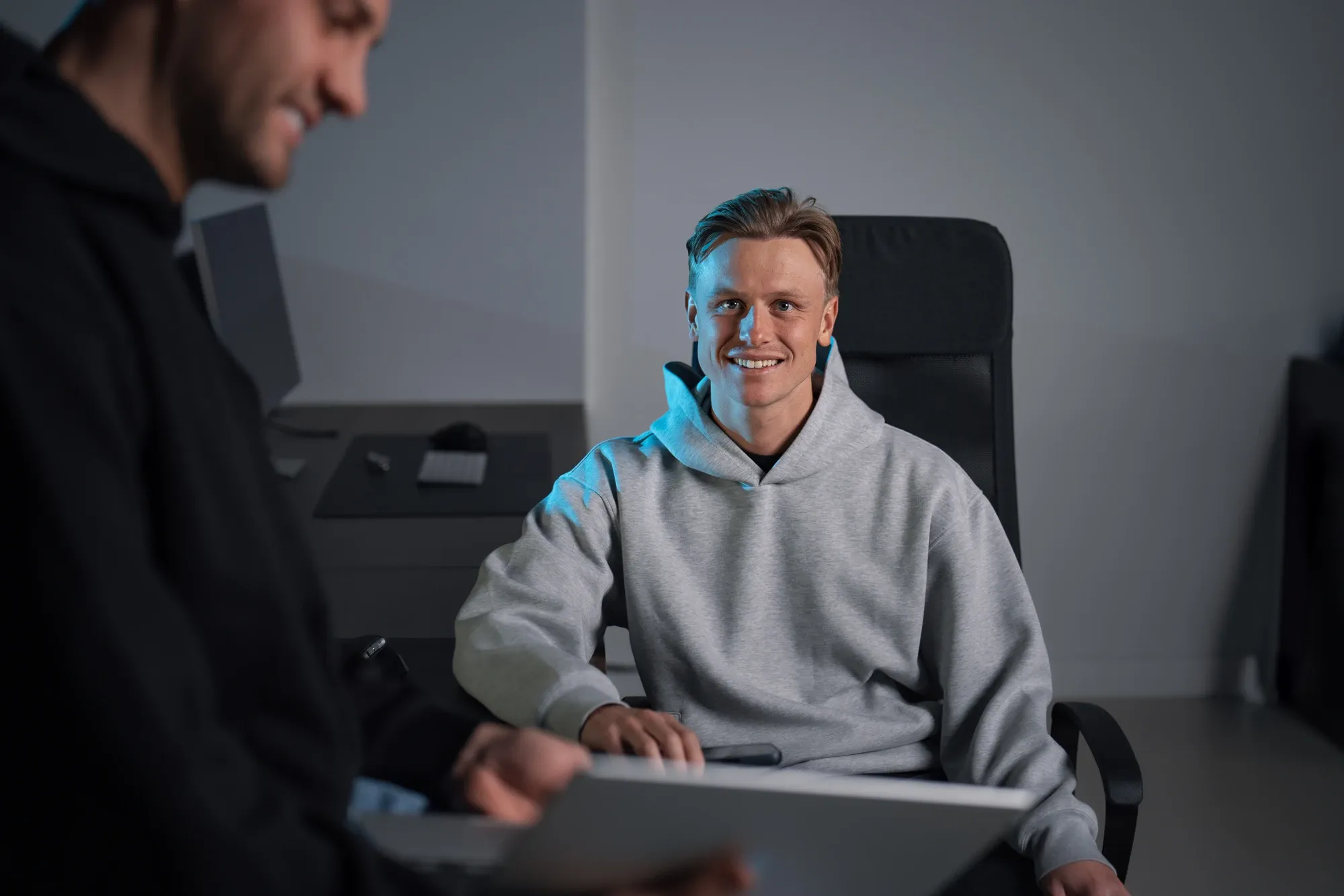 Collega van Klubb in een grijze hoodie lacht vanuit zijn bureaustoel, met een laptop op de voorgrond