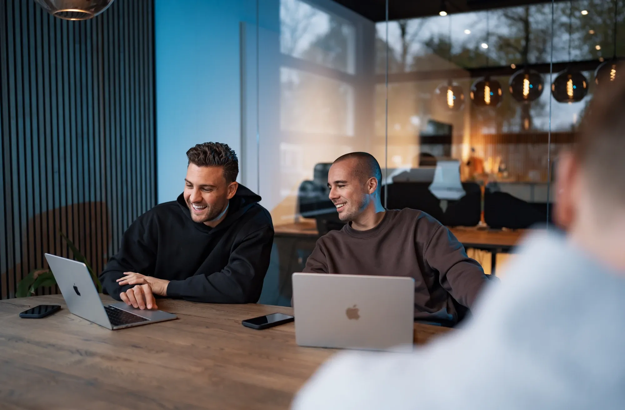 Twee collega’s van Klubb lachen aan een houten tafel met laptops, bij het raam op kantoor