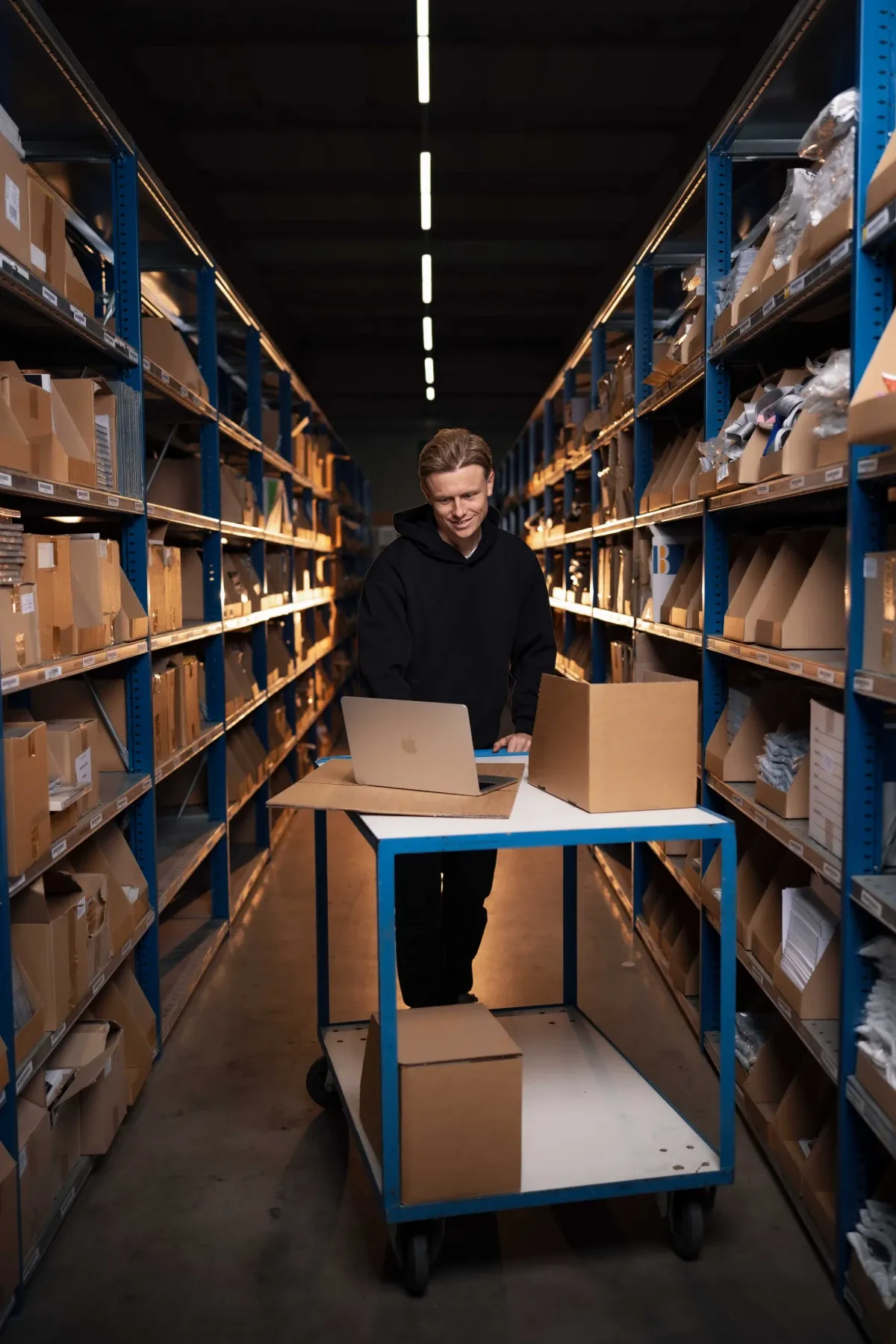 Specialist van Klubb achter een laptop op een rolwagen, tussen de stellingen van een magazijn