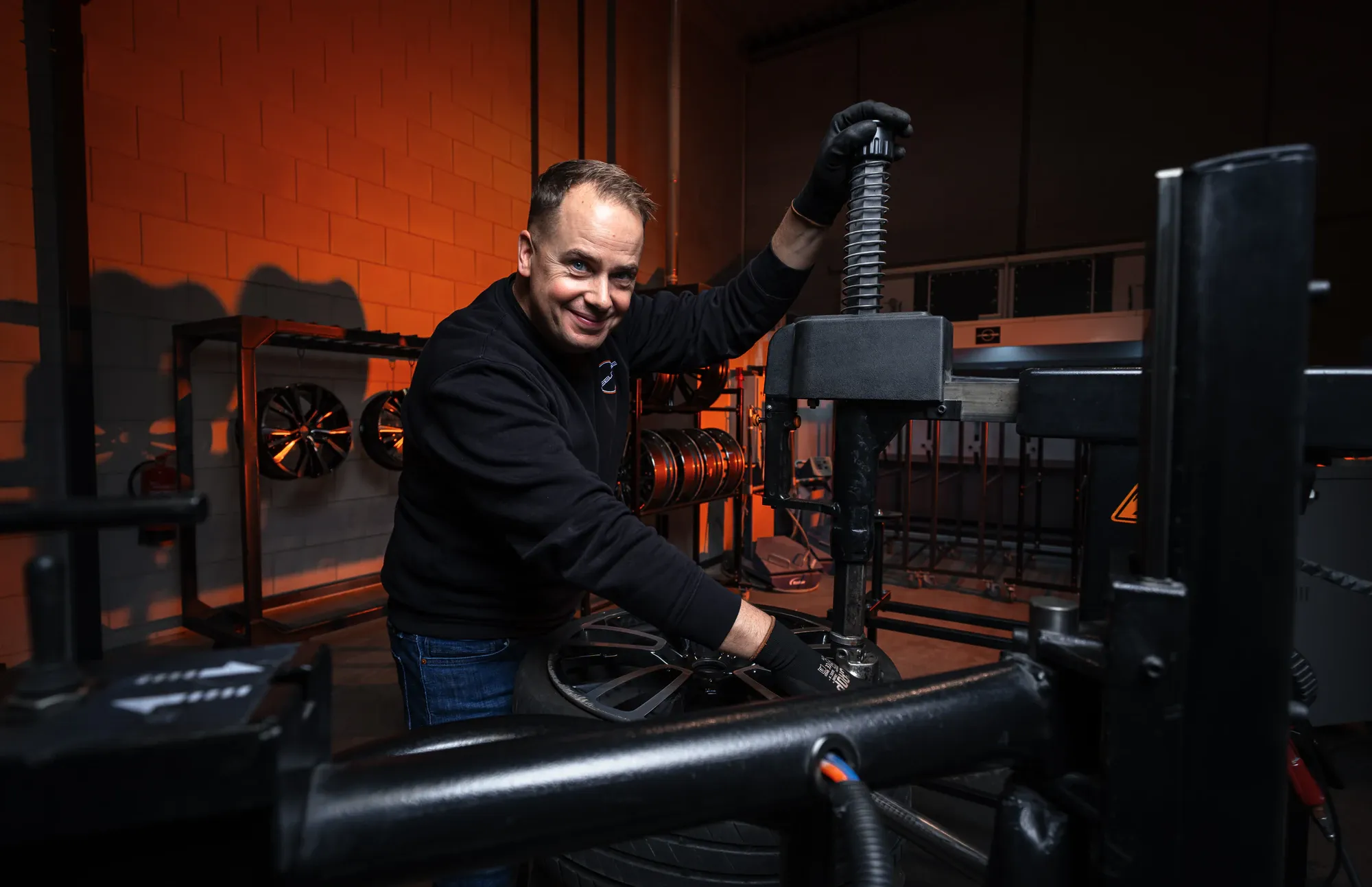 Lachende medewerker van Wheelness aan de bandenmontagemachine, met velgen in een rek tegen de oranje verlichte wand