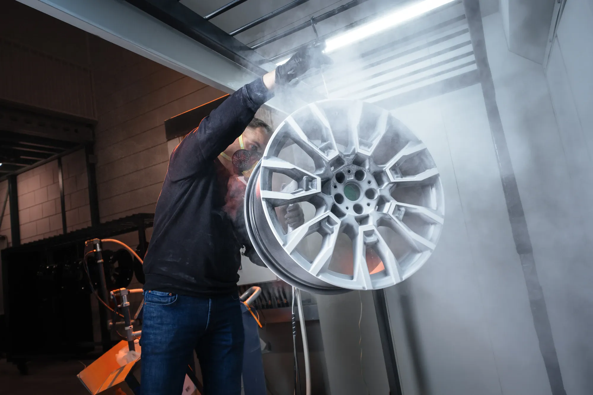 Medewerker van Wheelness met een stofmasker spuit poeder op een zilveren velg in de cabine